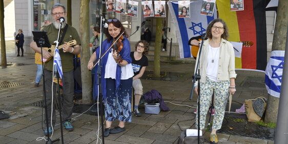 Eröffnung durch Rosi Dicke (Freunde Israels). Im Hintergrund Bilder von israelischen Kindern und Jugendlichen, die noch in Geiselhaft sind. Links: Birte Lindtstaedt, Jens Brettschneider, Cornelia Ziemke und die Gitarristin Olga, die an diesem Tag für die Musik zuständig waren. (Foto: Kanneberg)